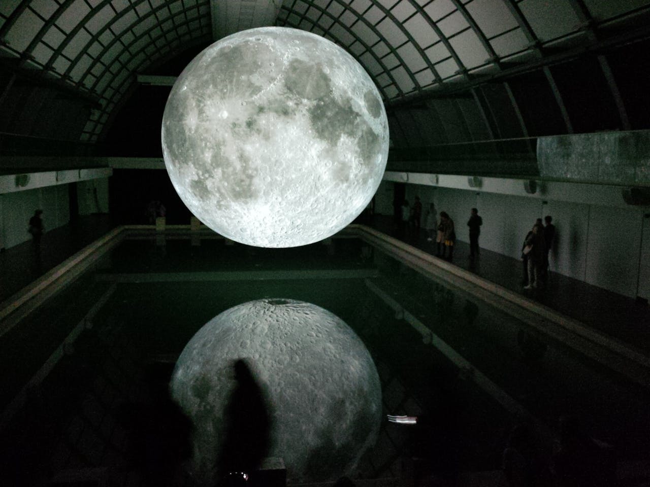 Mesmerizing moon installation reflected in water indoors with people observing.